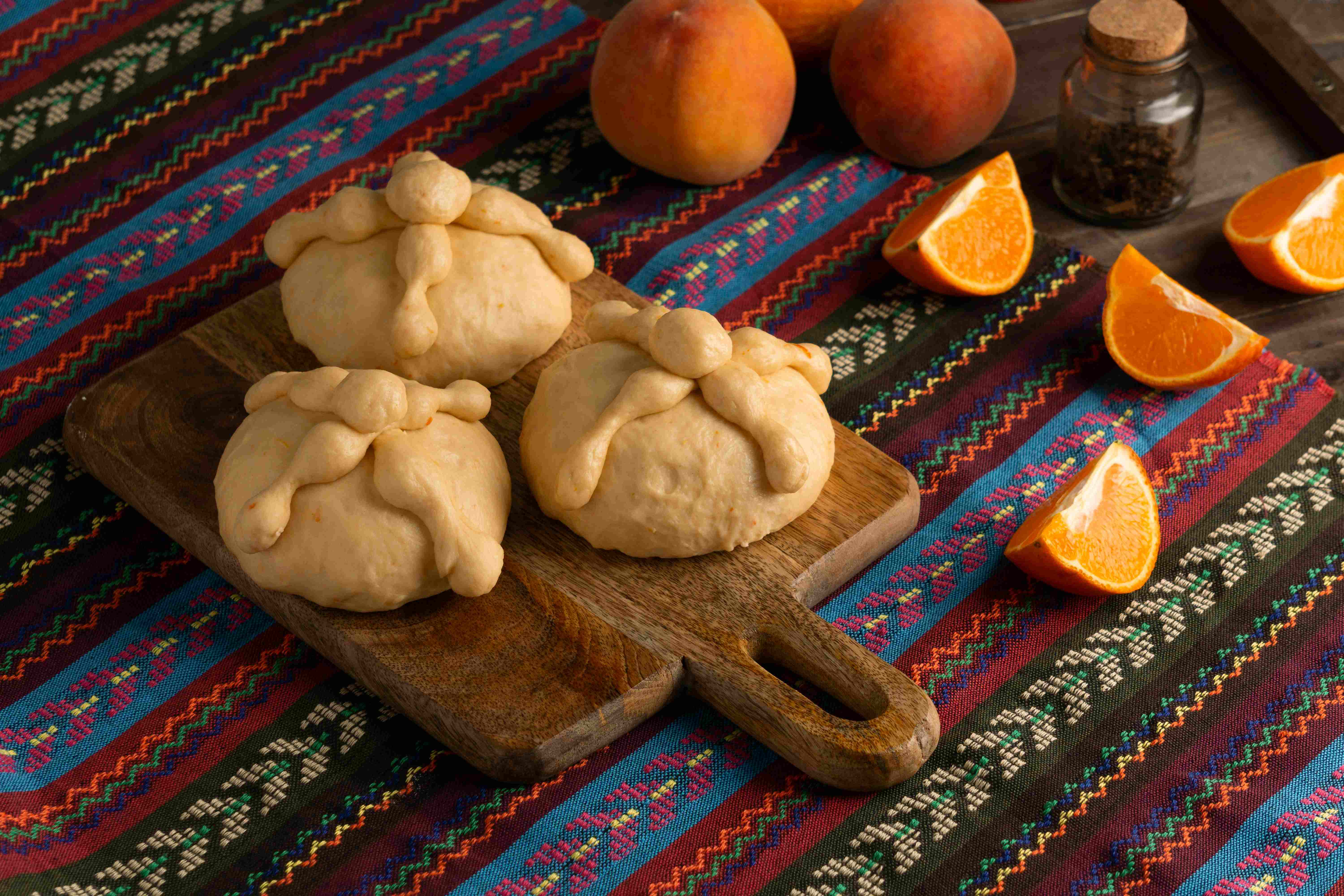 3 panes de muerto sobre una tabla encima de un reboso de colores y naranjas cortadas en gajos a su lado