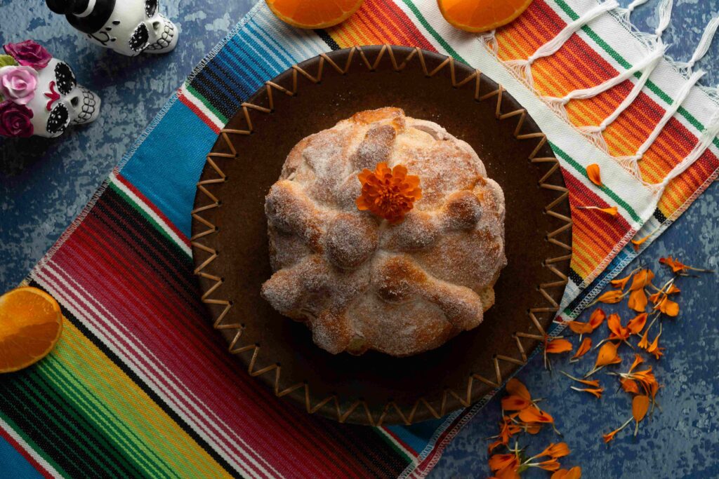 Pan de muerto sobre un reboso de colores y con una flor de cempasúchil encima de la azúcar