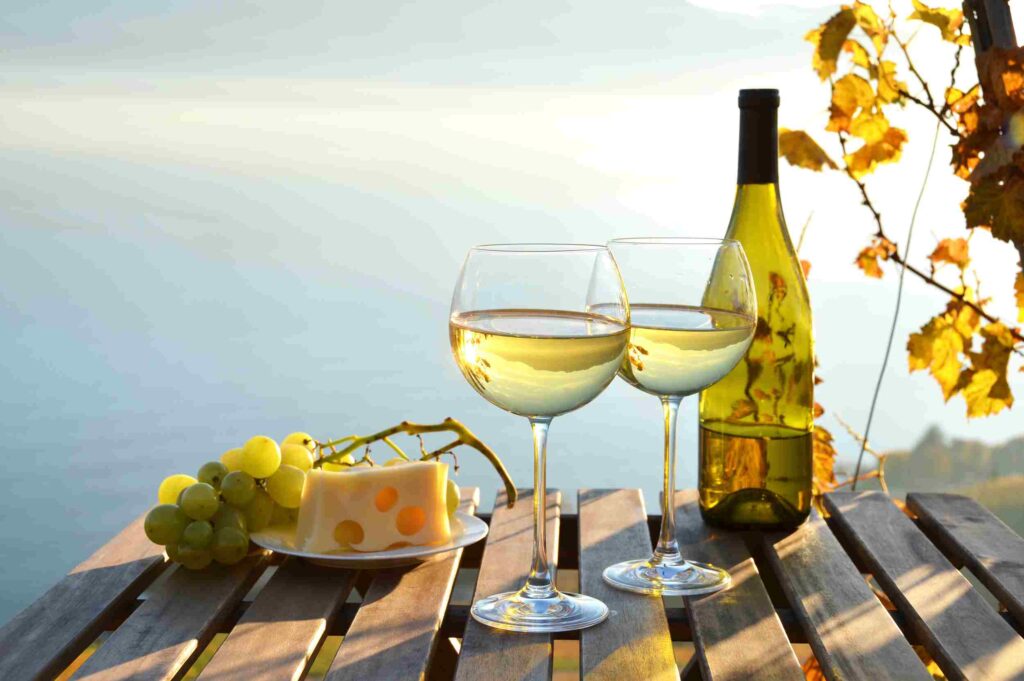 Dos copas de vino al lado de una botella, sobre una mesa, con queso y uvas, frente al mar